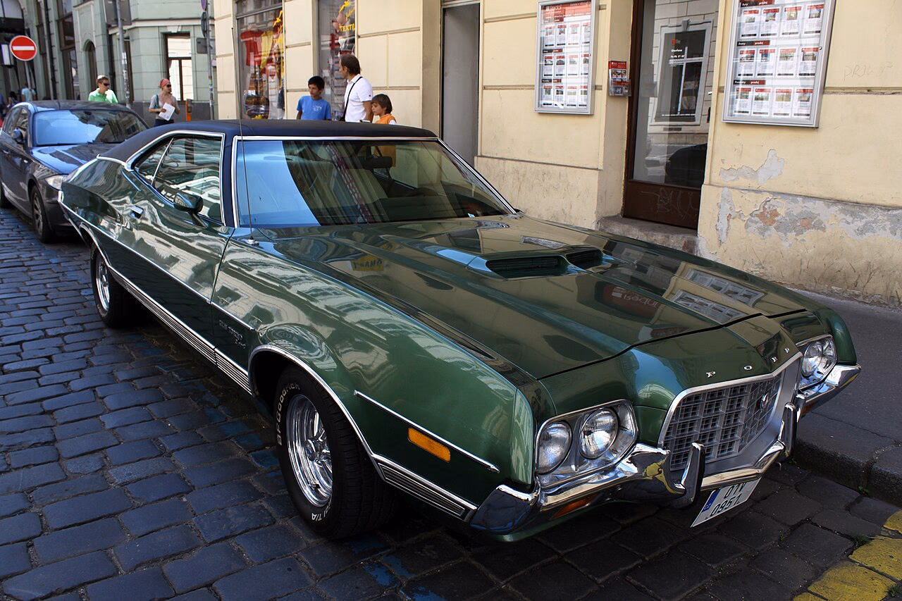 Grüner Ford Gran Torino, schräg von vorn auf einer gepflasterten Straße
